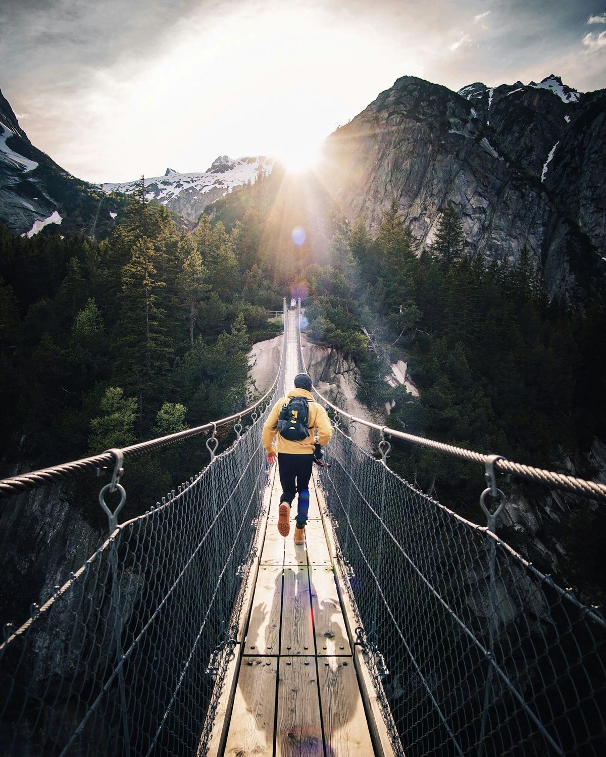 A hiker crossing a suspension bridge toward sunlit mountains